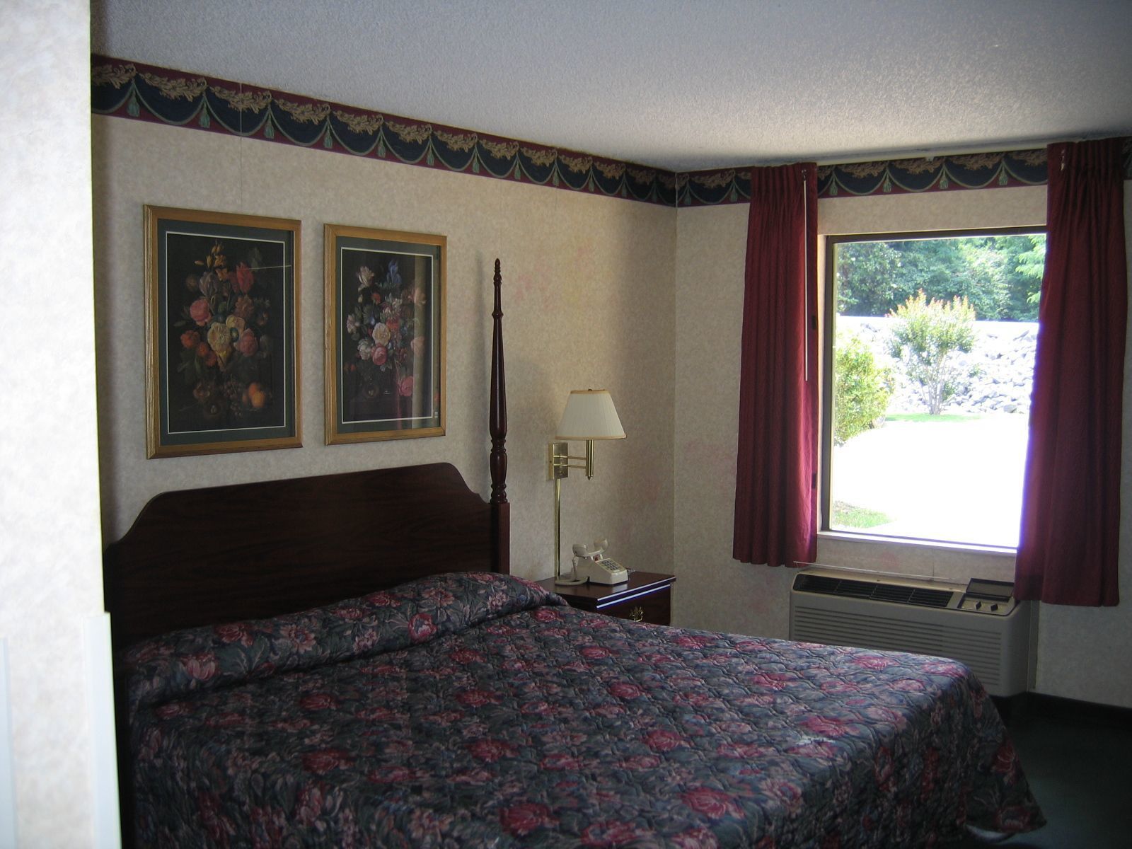 A hotel room with a bed, two framed floral paintings, a nightstand with a lamp, and a window with curtains.