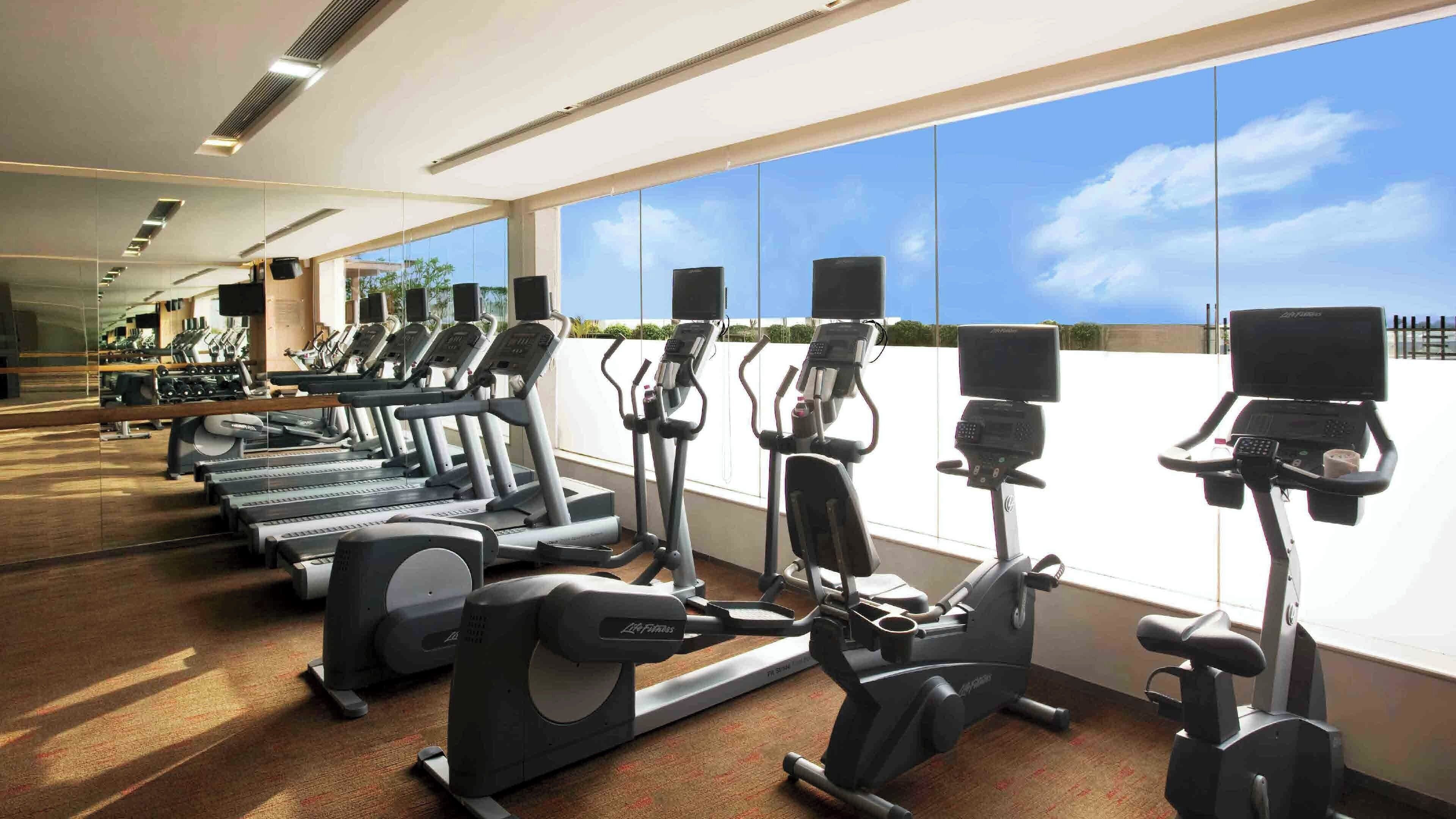 A gym with treadmills, elliptical machines, and exercise bikes, with large windows offering a view of the sky and clouds.