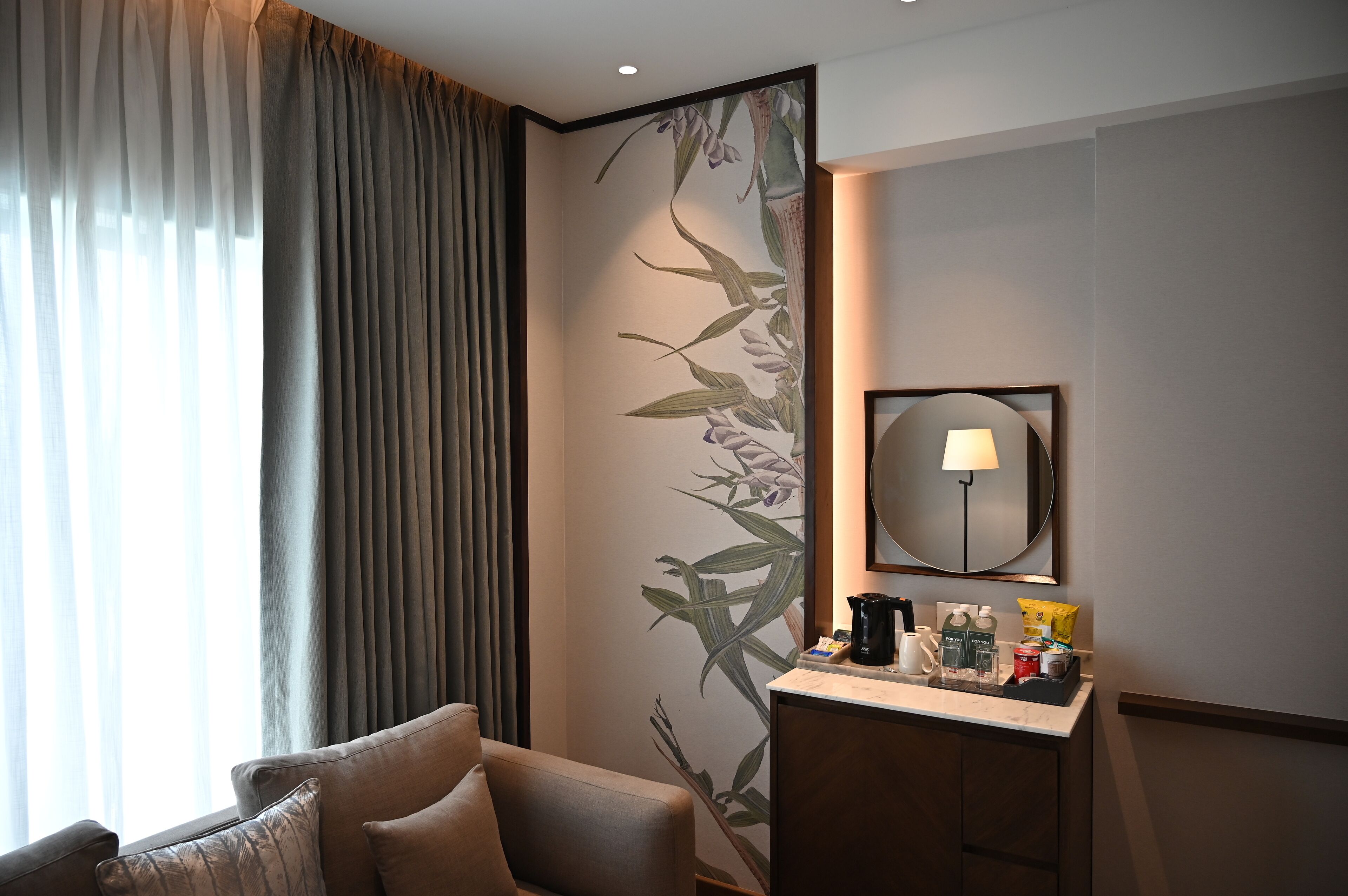 Une chambre d’hôtel avec un canapé, une table de chevet avec des articles de toilette, un miroir fixé au mur et une fenêtre avec des rideaux.