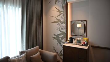 A hotel room with a sofa, a bedside table with toiletries, a wall-mounted mirror, and a window with curtains.