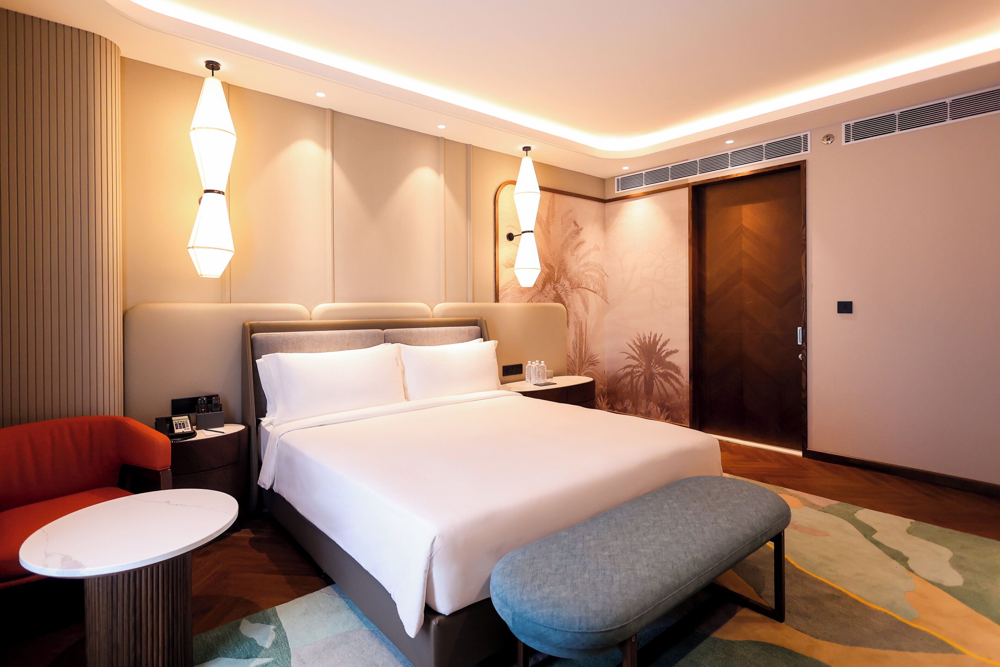 A hotel room with a large bed, a bedside table, a chair, a small table, a bench, and a wardrobe.