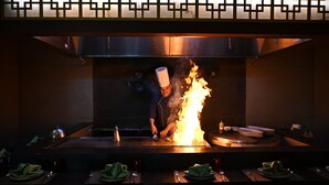 Uno chef cucina su una griglia con delle fiamme, in un ambiente da ristorante.