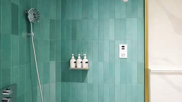 A bathroom with teal tiles, a showerhead, a shelf with toiletries, and a thermostat.