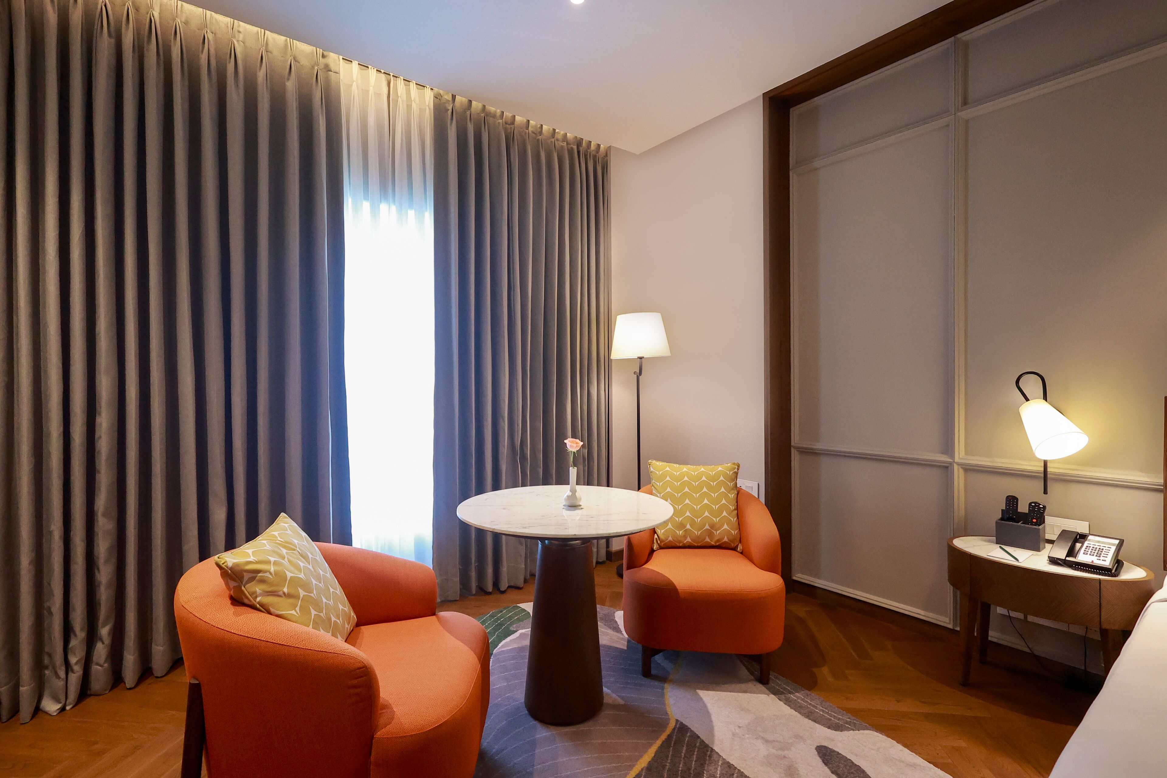 A hotel room with two orange armchairs, a round table with a vase, a lamp, and a phone on a side table.