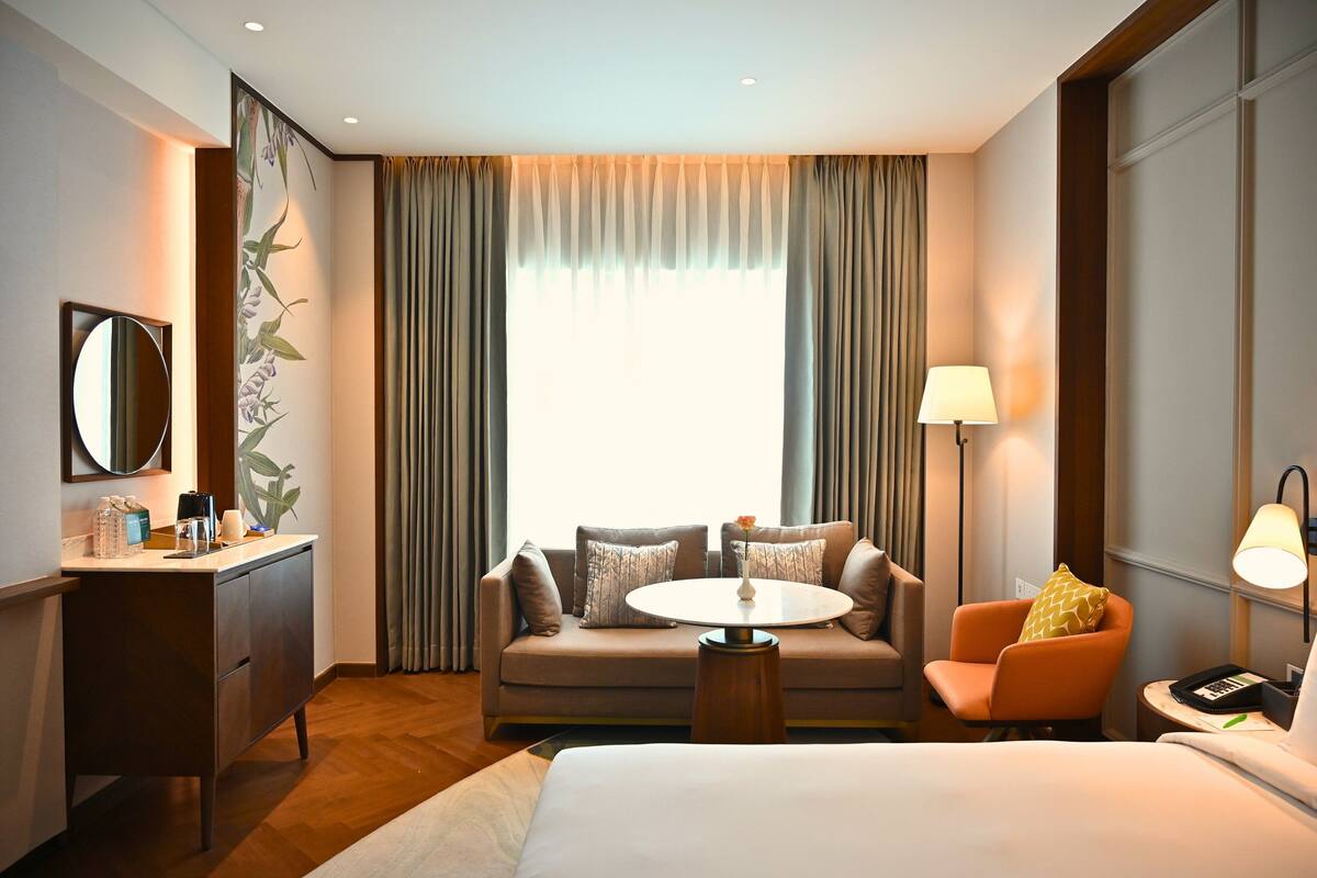 A hotel room with a bed, a sofa, a small table, a chair, a desk with a mirror, and a window with curtains.
