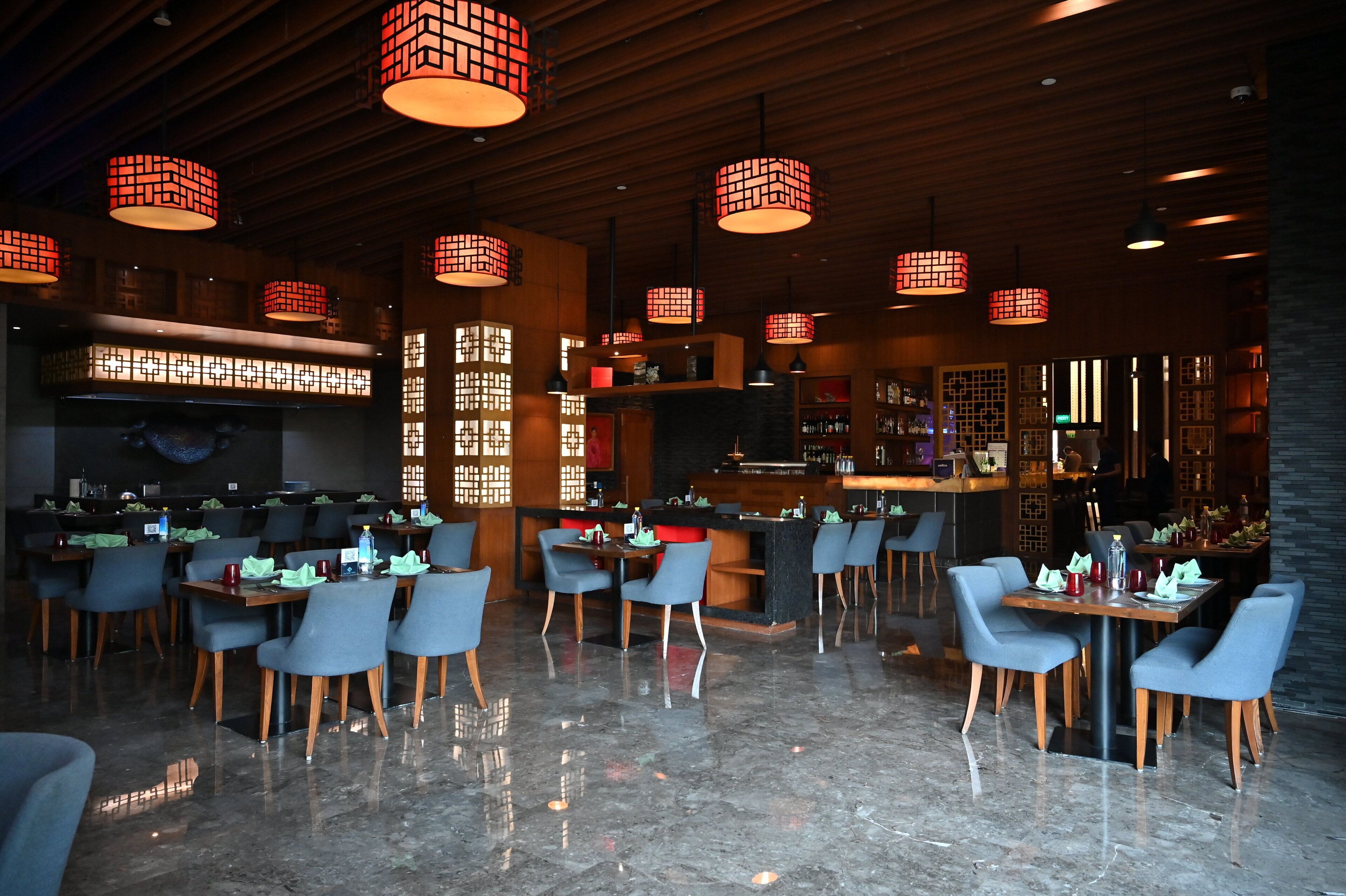 A modern restaurant with wooden ceiling panels, red hanging pendant lights, and a polished marble floor.