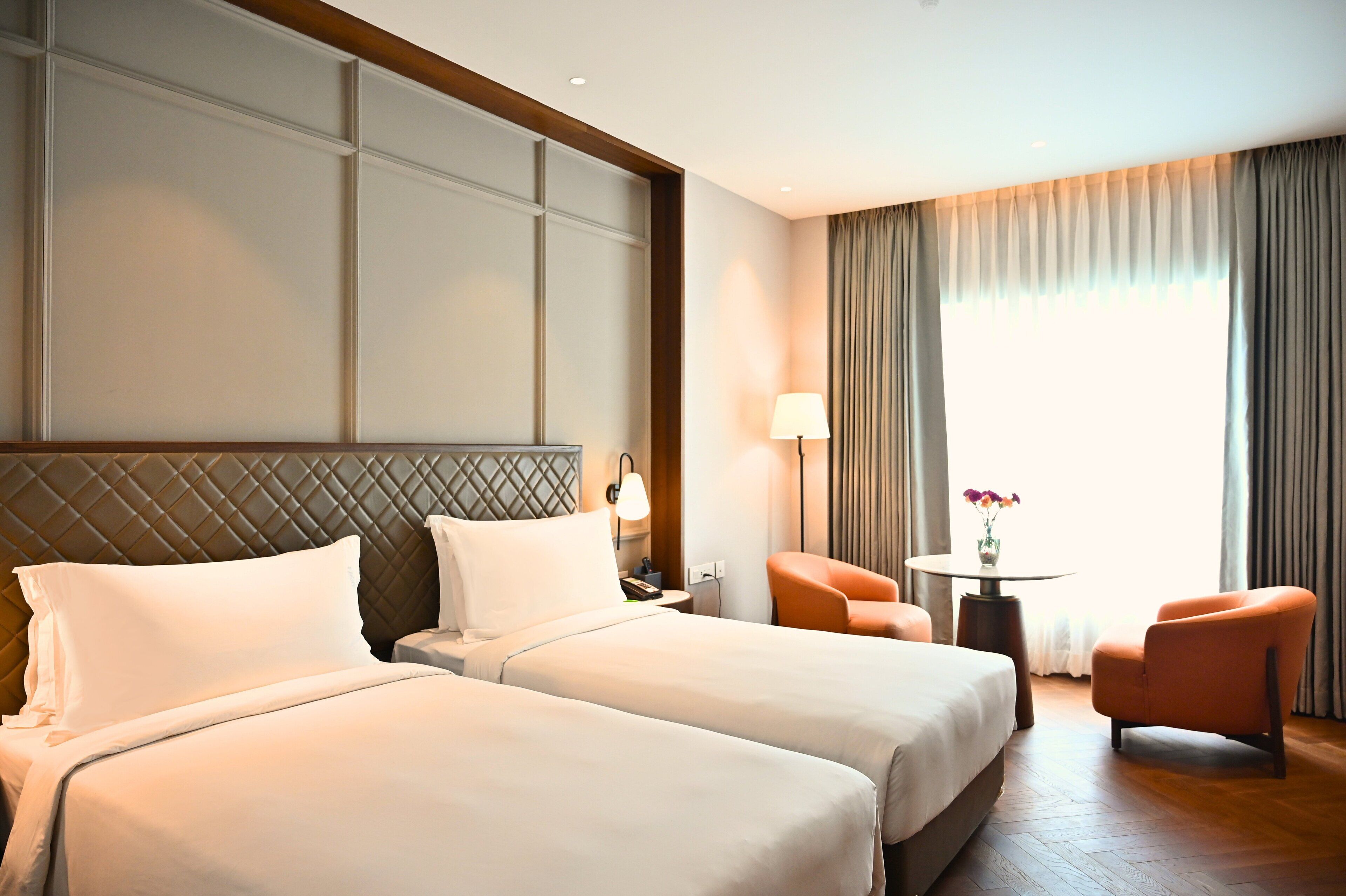 A hotel room with two beds, a nightstand, a lamp, a chair, and a window with curtains.