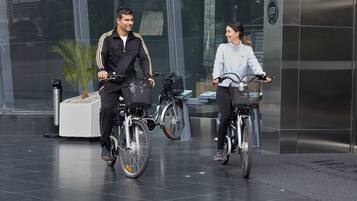 Two people riding bicycles on a tiled pavement in front of a modern building with a glass facade.