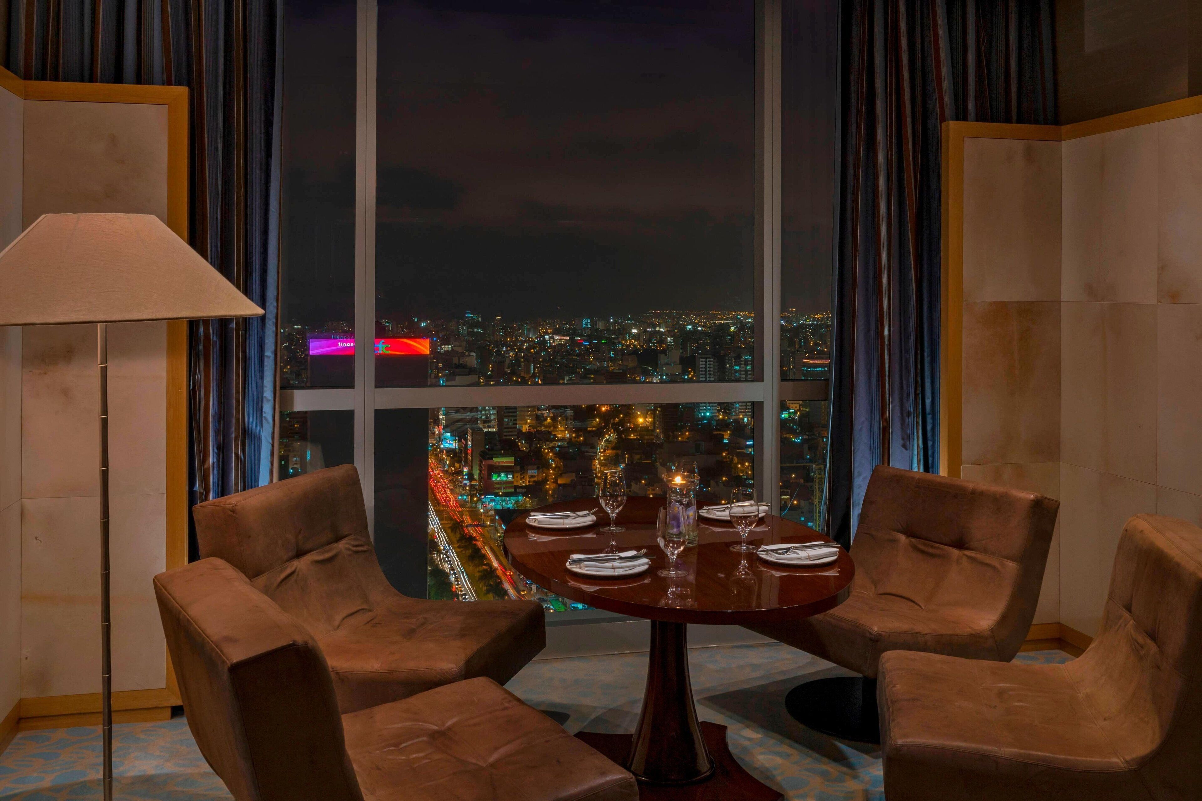 A dining area with a table set for four, two brown chairs, a lamp, and a view of a cityscape at night.