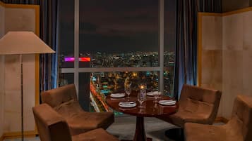 A dining area with a table set for four, two brown chairs, a lamp, and a view of a cityscape at night.