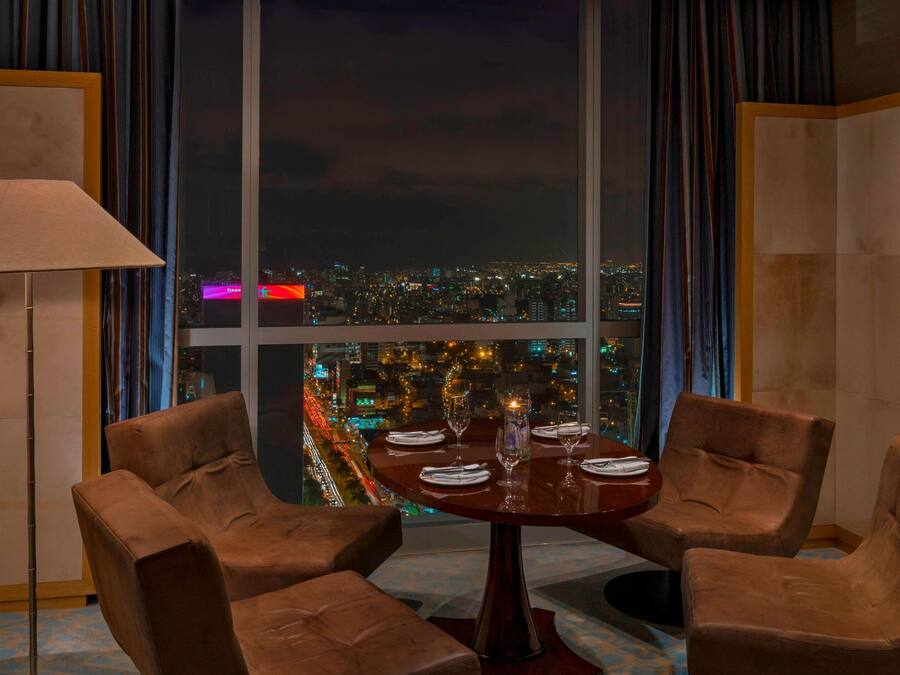 A dining area with a table set for four, two brown chairs, a lamp, and a view of a cityscape at night.