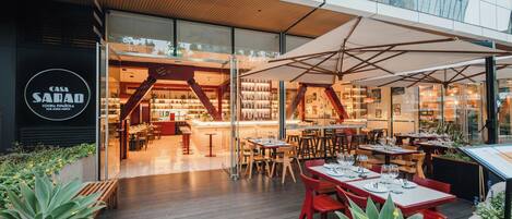 Un área de comedor exterior moderna con mesas y sillas, una terraza de madera y un restaurante con fachada de vidrio al fondo.