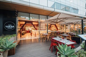 Un área de comedor exterior moderna con mesas y sillas, una terraza de madera y un restaurante con fachada de vidrio al fondo.
