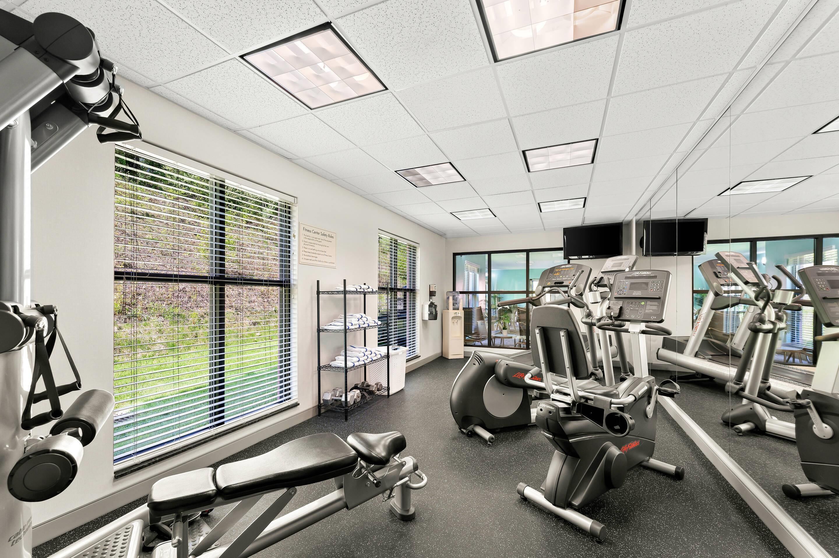 A gym with various exercise equipment, large windows with blinds, and a view of greenery outside.