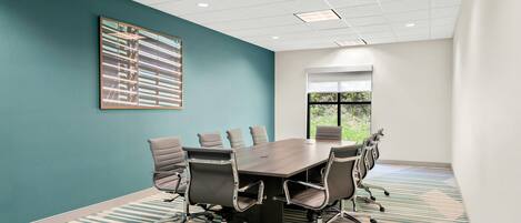 A conference room with a large table, six chairs, teal walls, a large window, and a ceiling with recessed lighting.