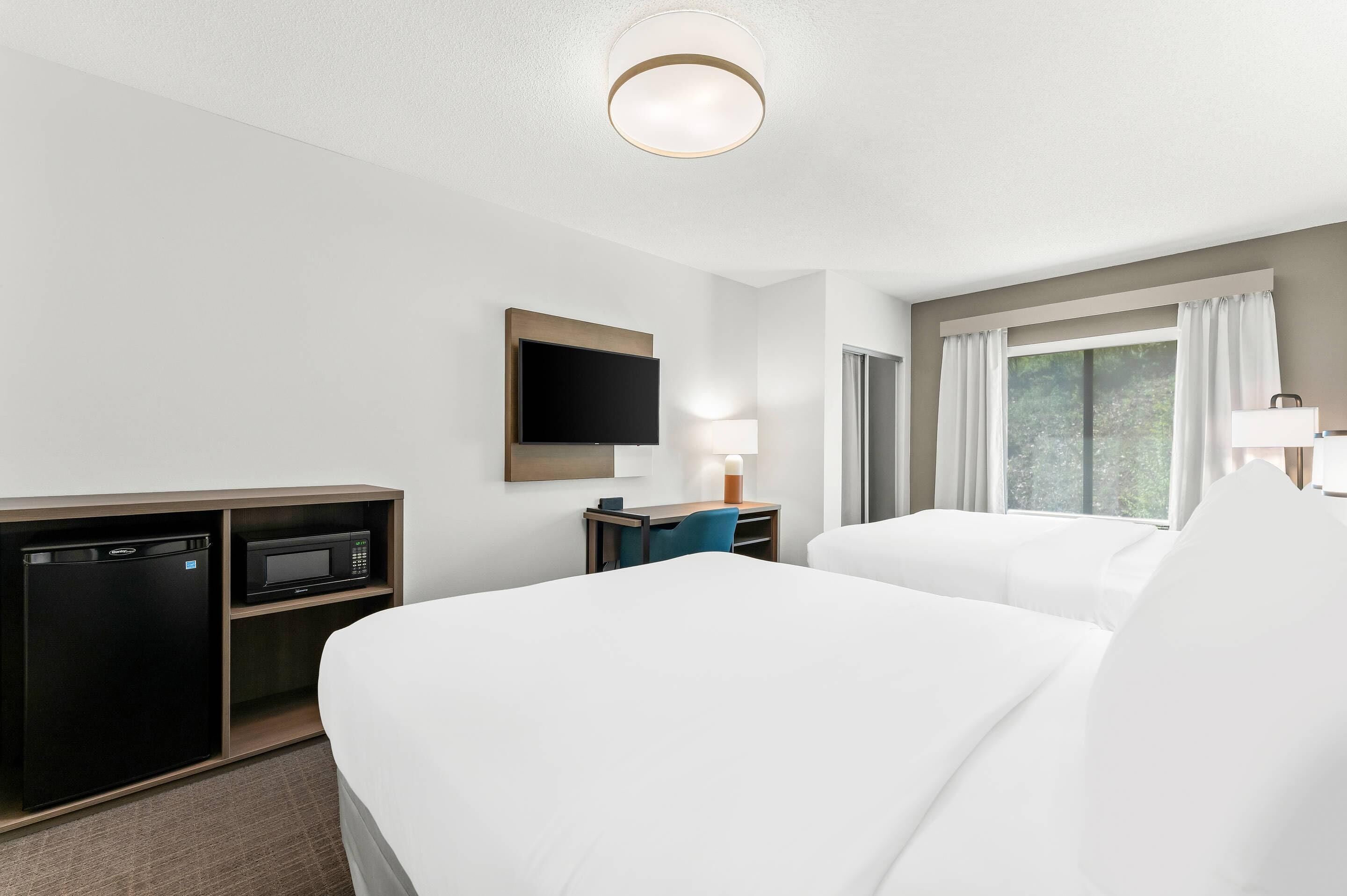 Hotelkamer met twee bedden, een bureau met stoel, een magnetron, een koelkast, een televisie en een raam met gordijnen.