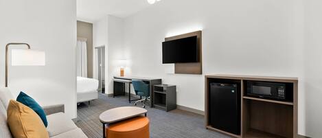 A modern hotel room with a sofa, a small table, a desk with a chair, a TV mounted on the wall, and a microwave.