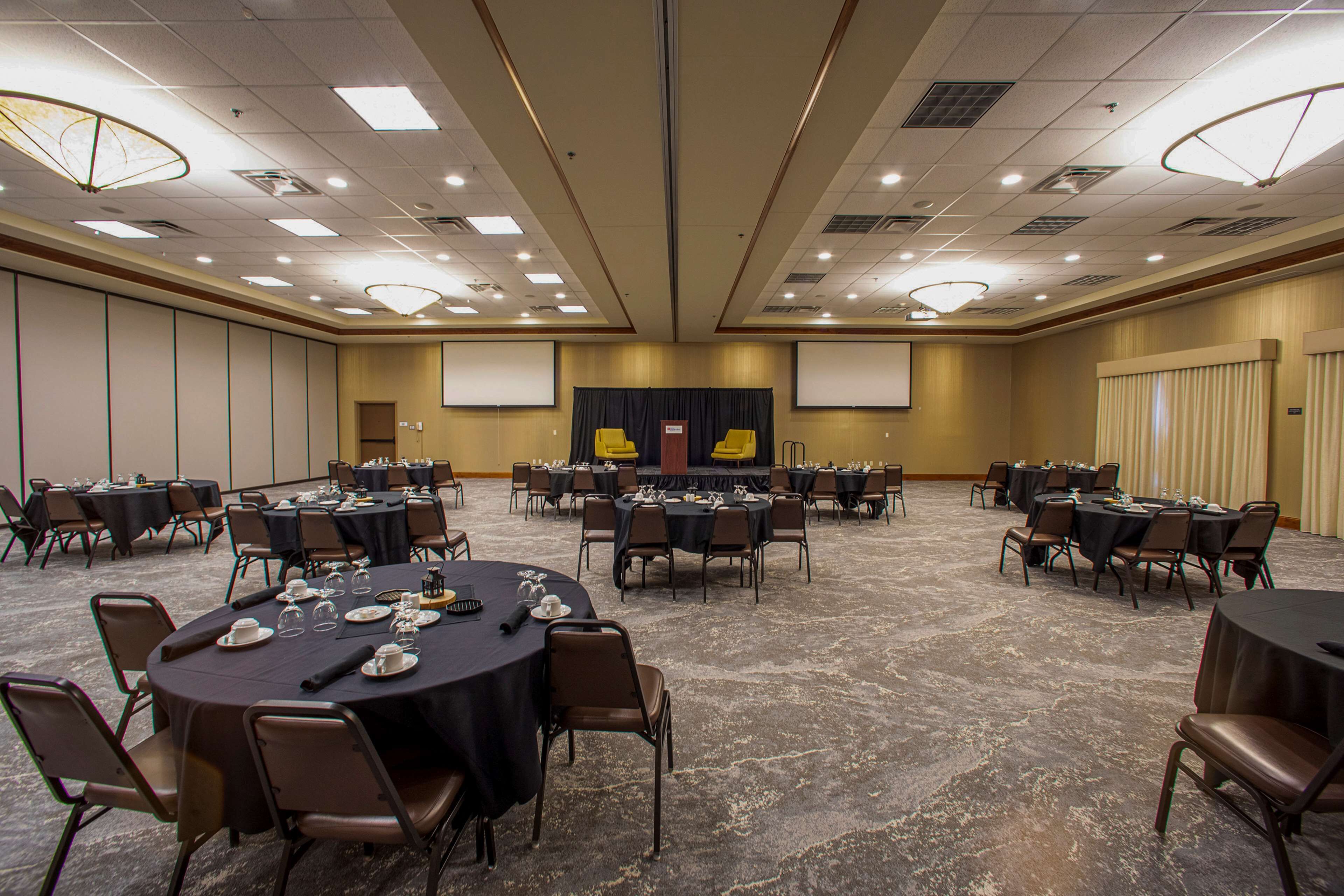 A large conference room with multiple round tables and chairs, equipped with presentation screens and a stage.
