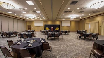A large conference room with multiple round tables and chairs, equipped with presentation screens and a stage.