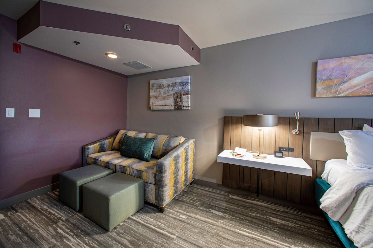 A hotel room with a sofa, ottoman, bedside table, lamp, and a bed.