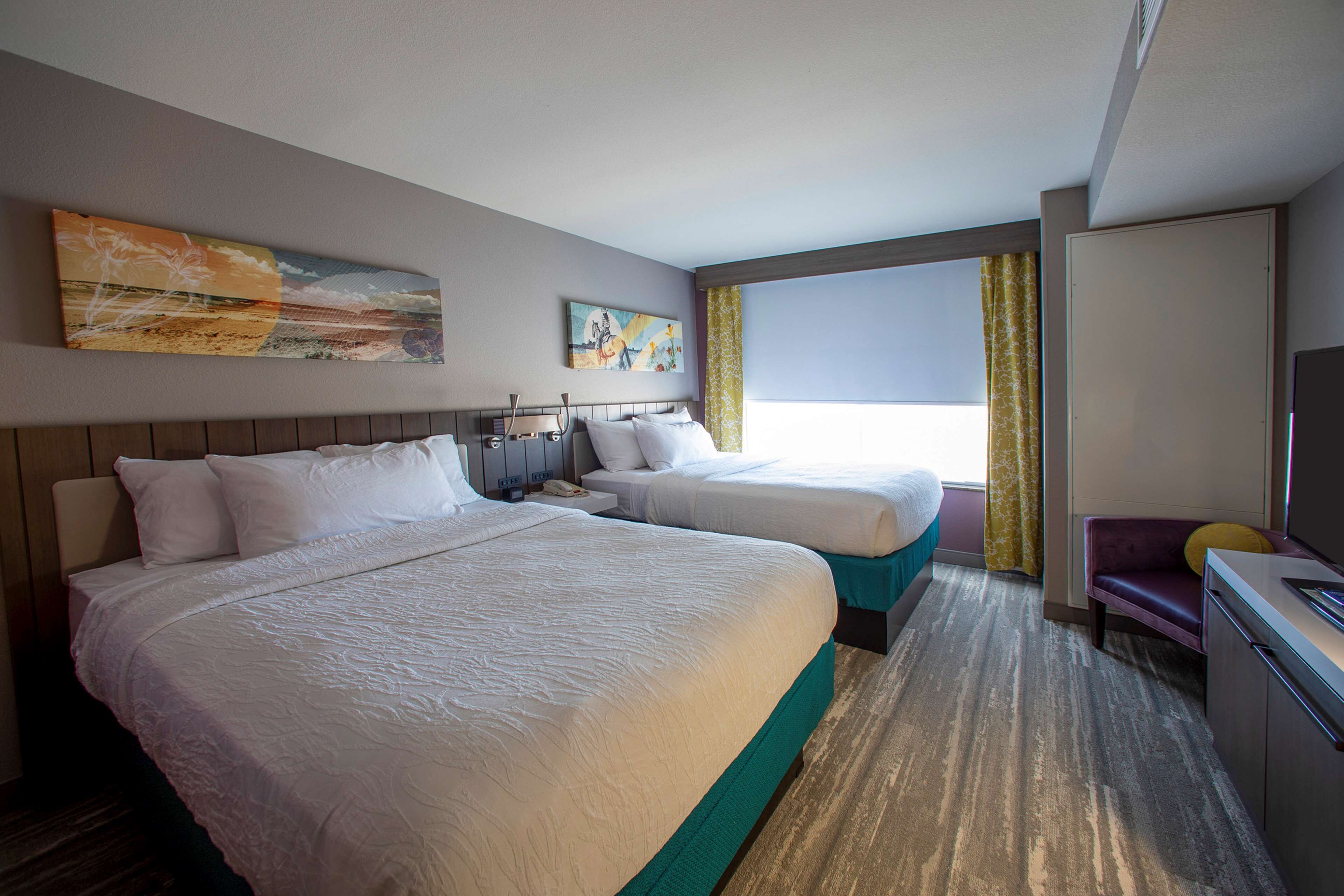 Ein Hotelzimmer mit zwei Betten, einem Schreibtisch, einem Fernseher und Bildern an den Wänden.