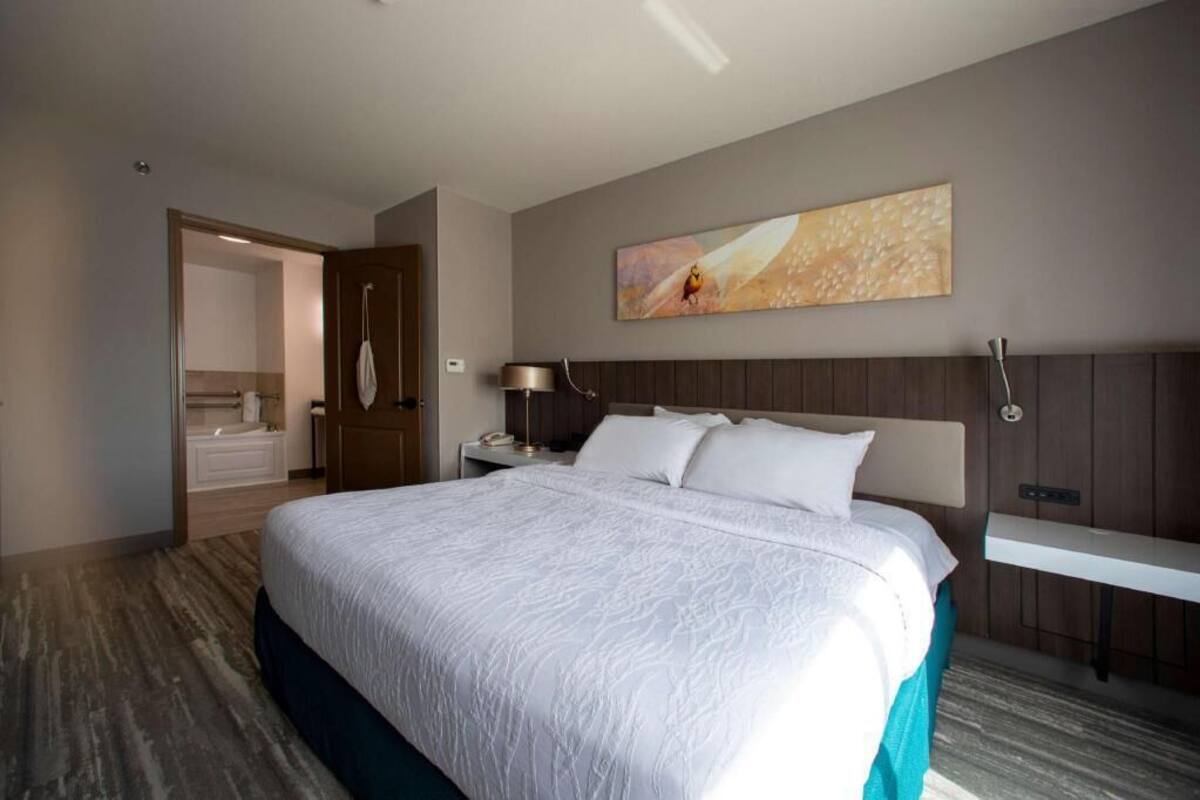 A hotel room with a large bed, a nightstand, a wall-mounted lamp, a painting, and a bathroom visible through an open door.