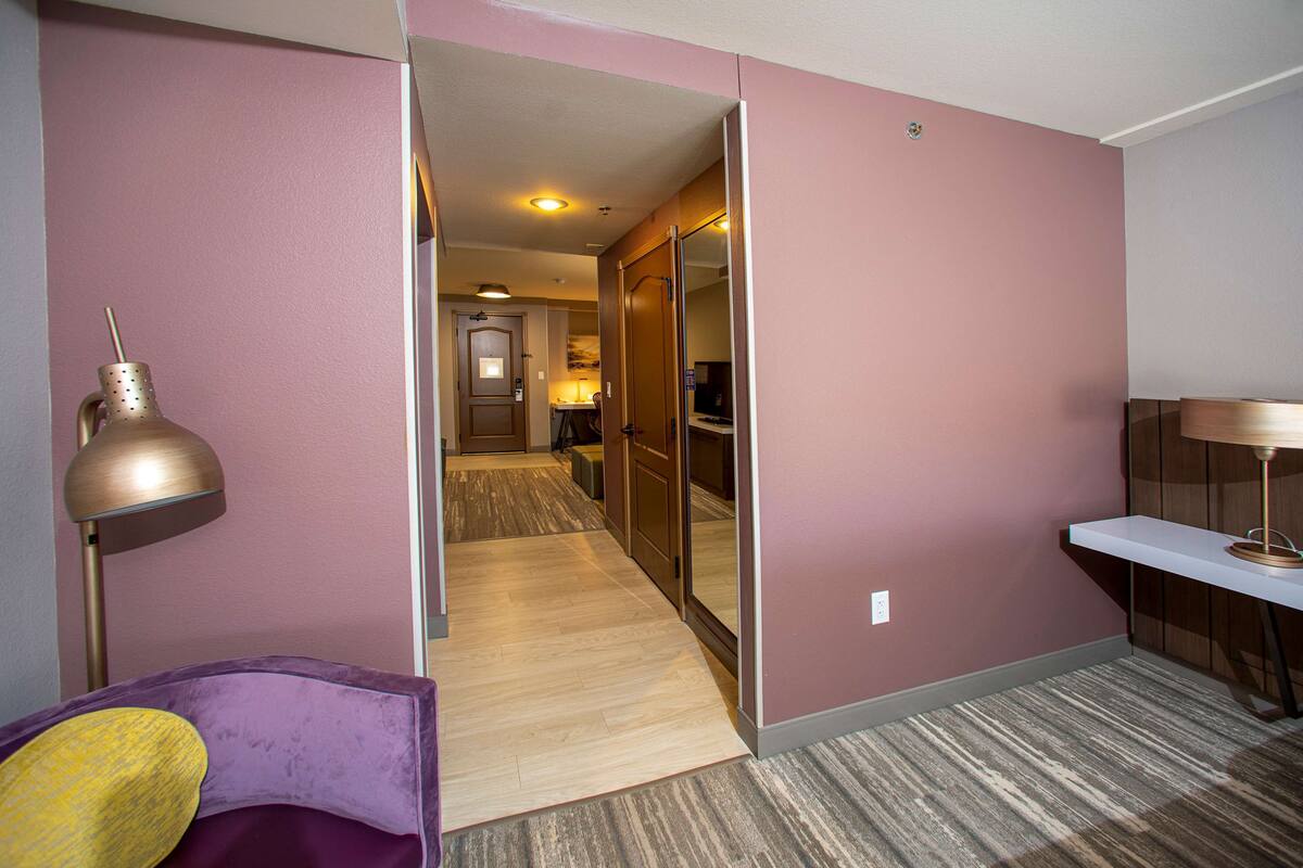 A hotel room with a purple armchair, a lamp, a wooden floor, and a hallway leading to a bathroom.