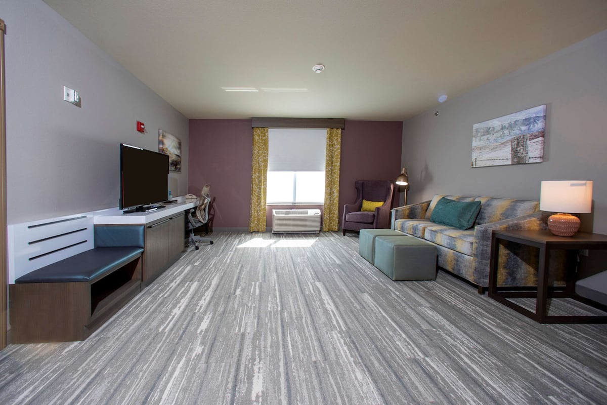 A compact hotel room with a flat-screen TV, a desk, a sofa, a small table with a lamp, and a window with curtains.