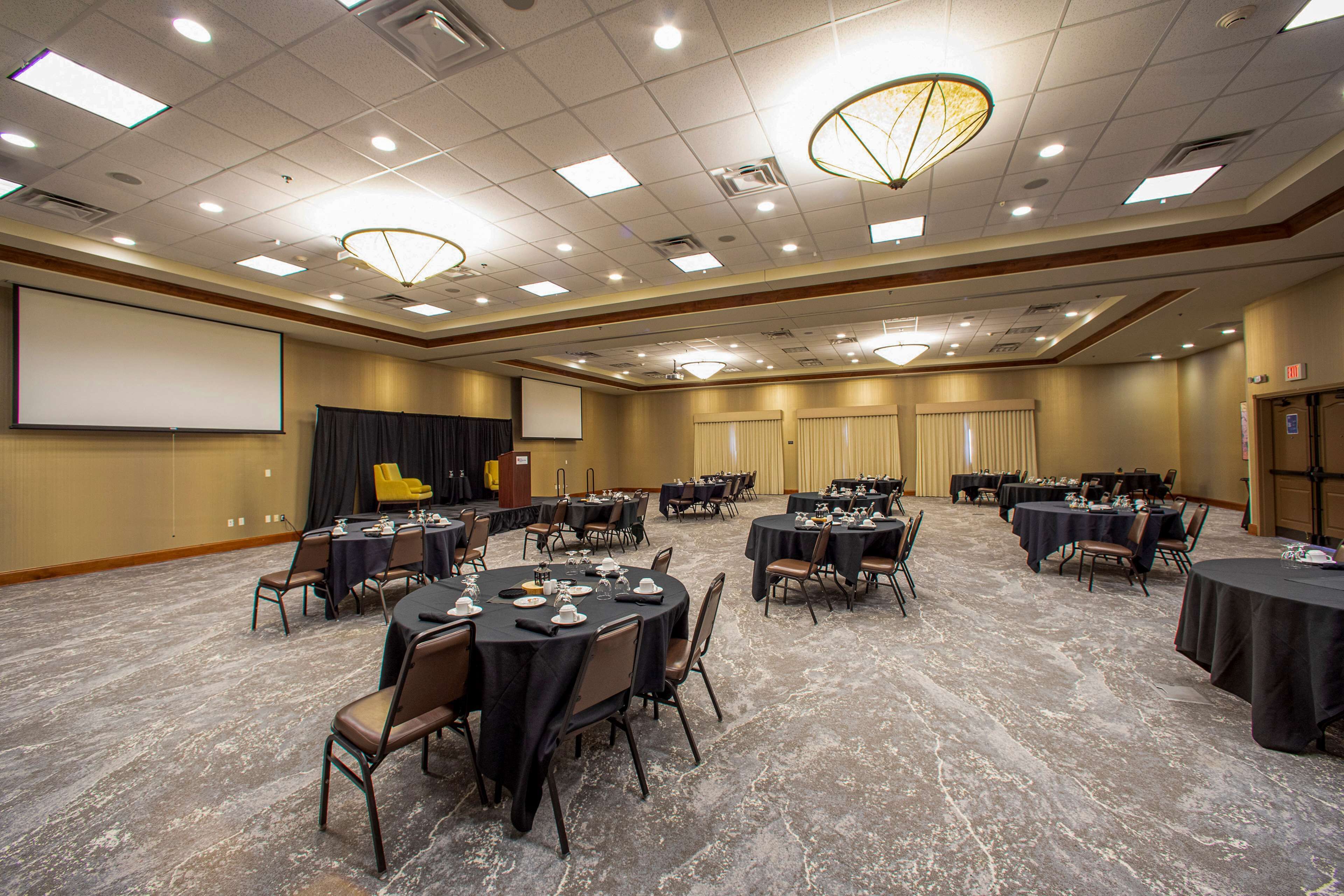 A conference room with multiple tables and chairs, a projector screen, and a stage with curtains.