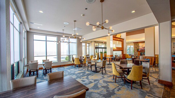 Een ruime hotel lobby met meerdere eettafels en stoelen, grote ramen en een kamerbreed tapijt met patroon.