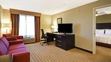 A hotel room with a bed, a desk with a computer, a TV, a red sofa, and a window with curtains.