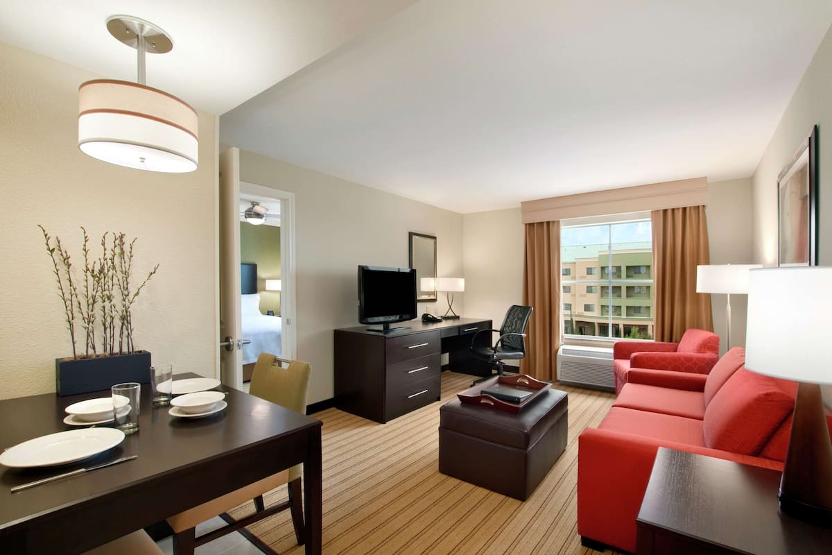 A hotel room with a dining table set, a TV, a sofa, and a window with curtains.