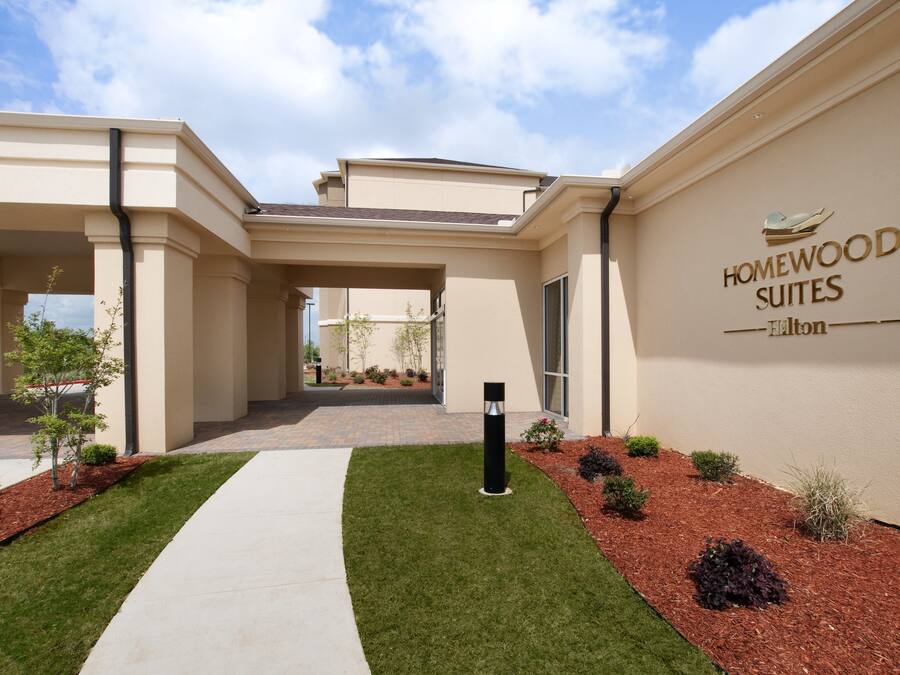Entrada de un hotel con un camino despejado hacia el edificio principal, un cartel que dice 'Homewood Suites Hilton' y un área ajardinada con arbustos pequeños y césped.