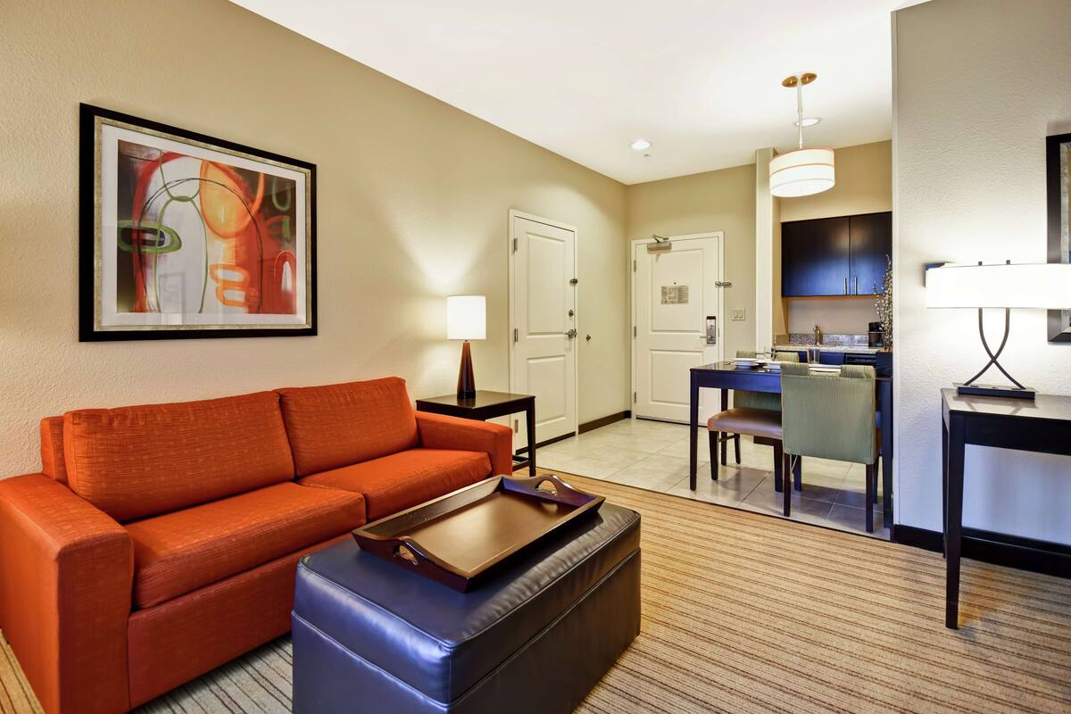 A hotel room with a sofa, ottoman, framed artwork, a desk, chair, and a kitchen area.