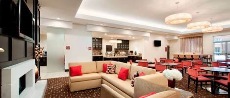 A modern hotel lobby with a sectional sofa, dining area, kitchen, and a fireplace.