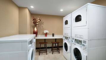 A laundry room with stacked washing machines, a vase with dried flowers, and two wooden stools.