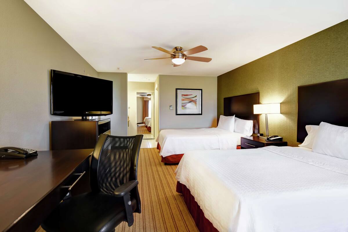A hotel room with two beds, a desk with a chair, a television, and a ceiling fan.
