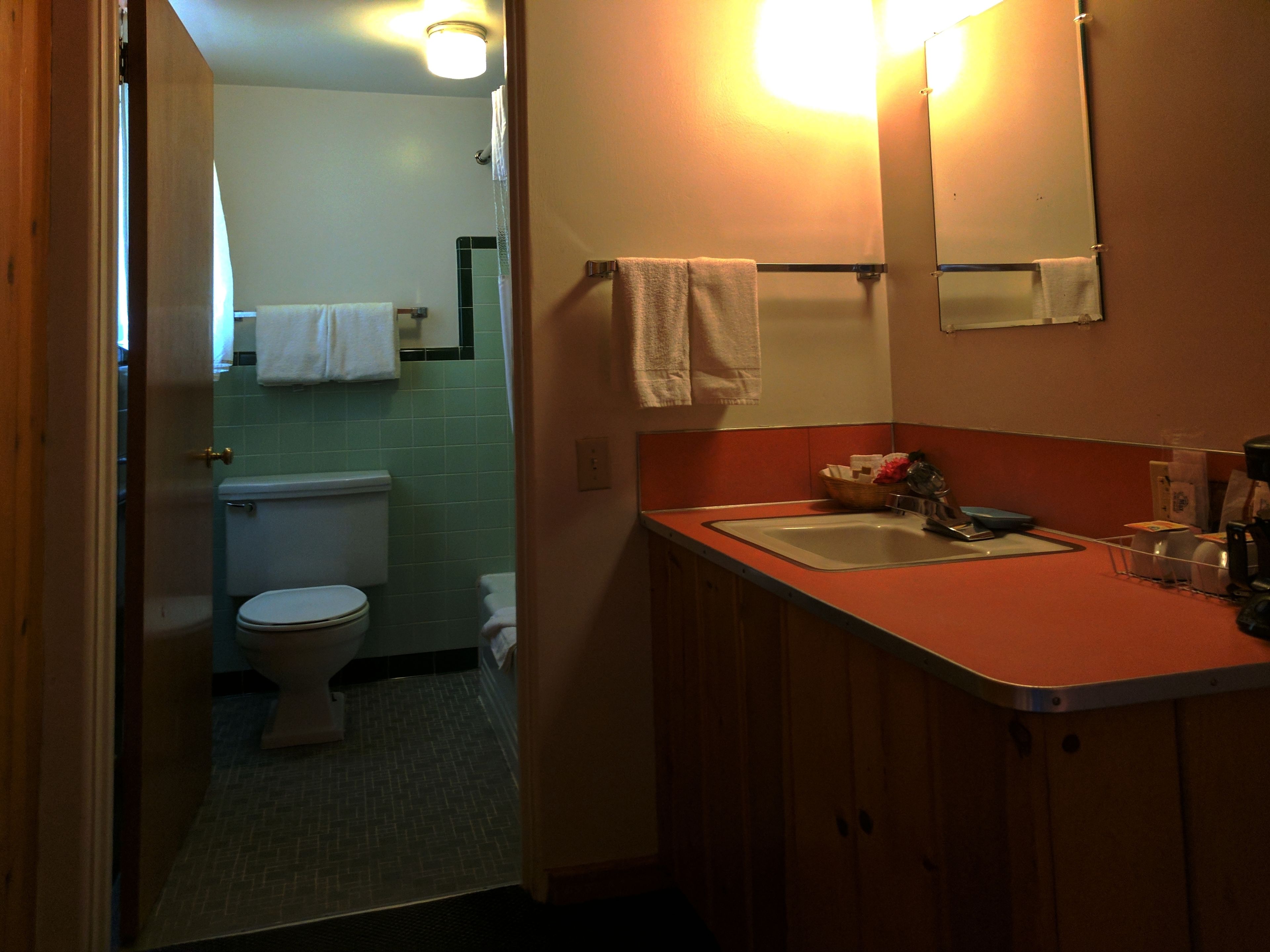 A bathroom with a toilet, sink, and mirror. There is a towel rack above the sink and a basket on the counter. The walls are tiled, and there is a door leading to another room.