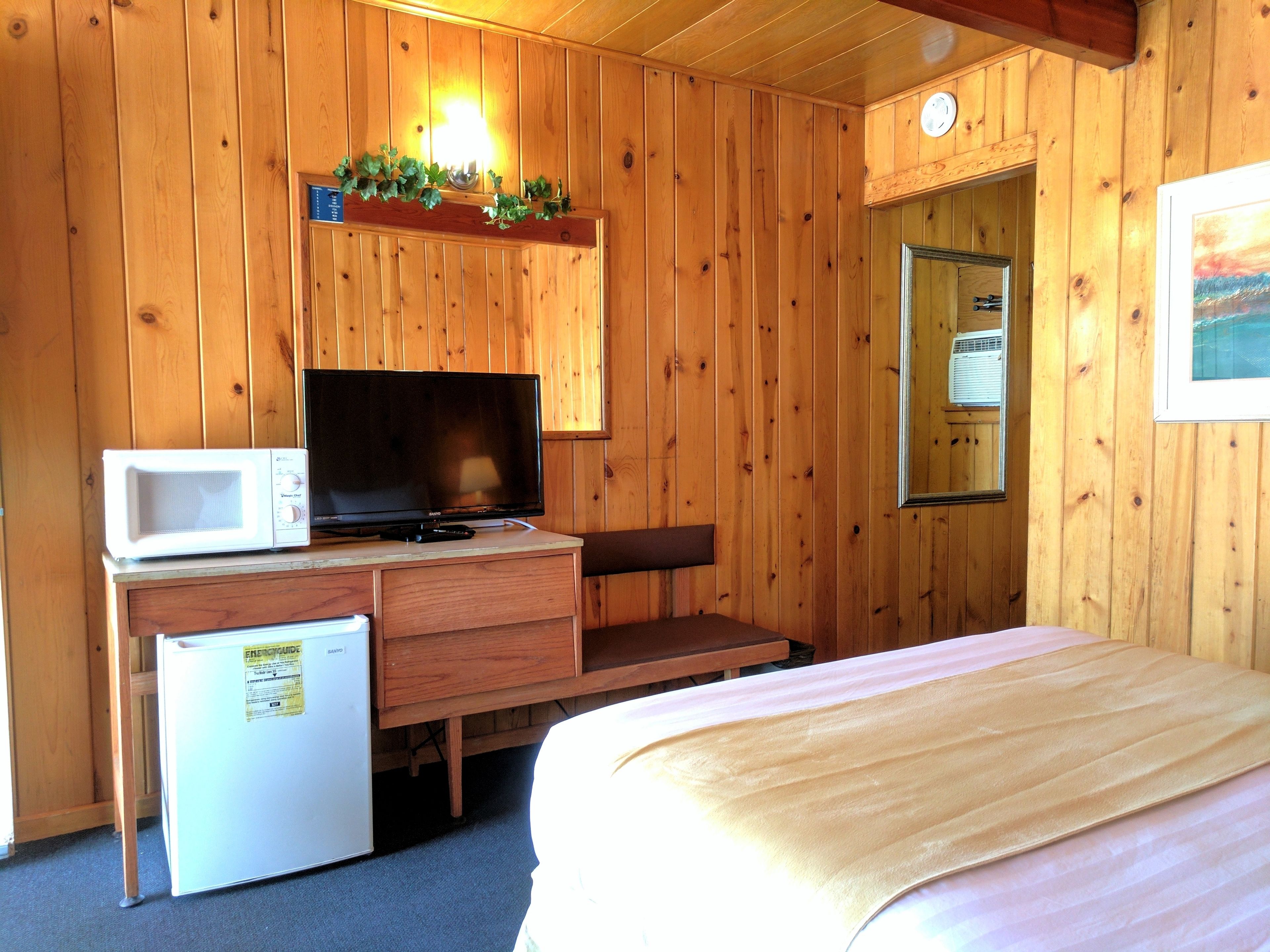 A wooden cabin room with a bed, a desk with a television, a microwave, and a refrigerator.