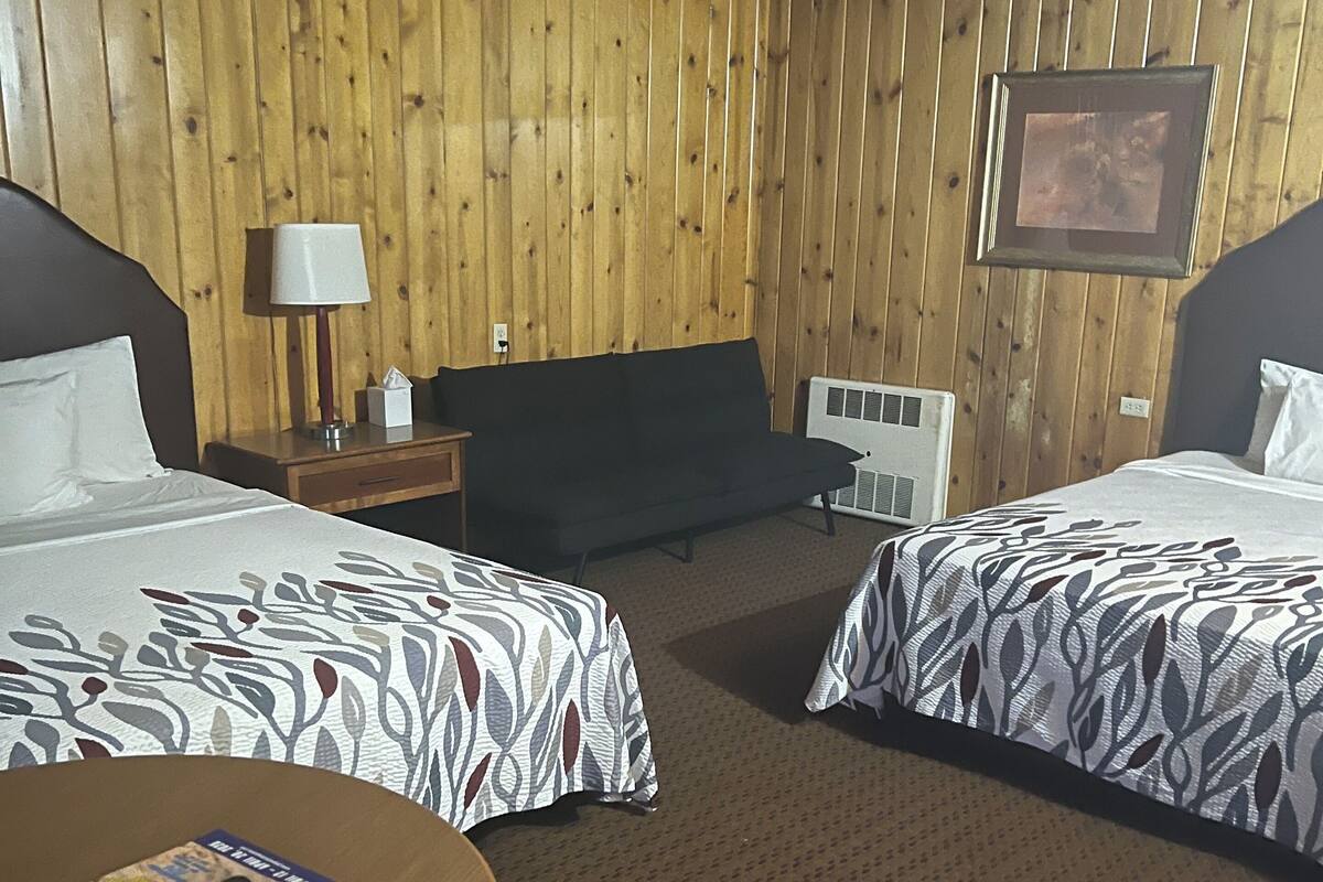 Ein Hotelzimmer mit zwei Betten, einer Holzwand, einem gerahmten Bild, einem Nachttisch mit Lampe und einem Tisch mit einer Zeitschrift.