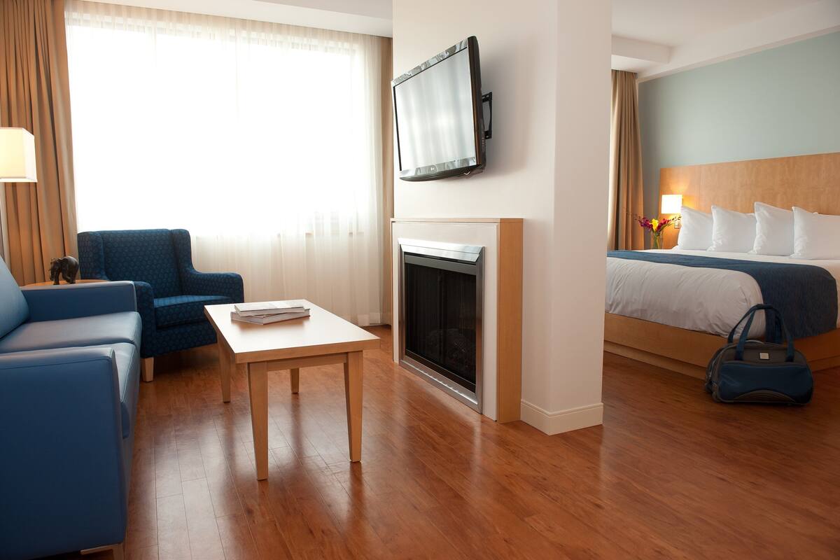 A hotel room with a sofa, a table, a fireplace, a TV mounted on the wall, and a bed with pillows.
