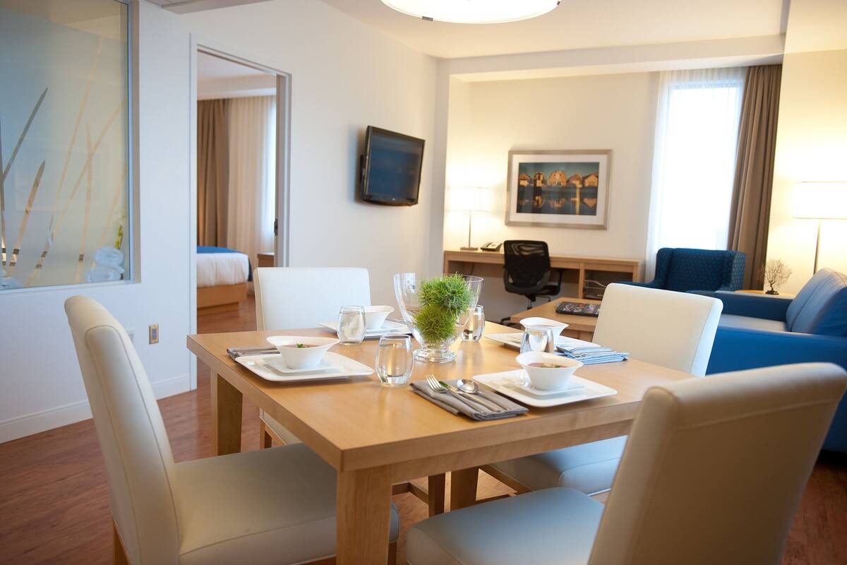 A hotel room with a dining table set for two, a blue sofa, a desk with a chair, and a bed in the background.