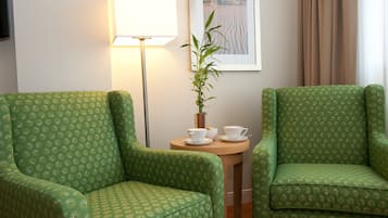 Two green patterned armchairs, a small wooden table with two white cups, a lamp, and a framed picture on the wall.