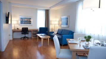 A hotel room with a wooden floor, a desk with a chair, a sofa, a dining table set for two, and a TV mounted on the wall.