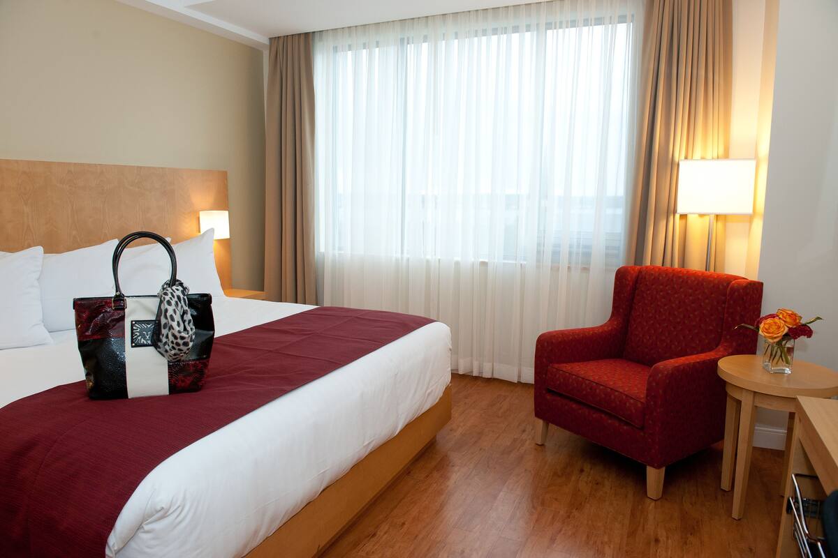 A hotel room with a bed, a red armchair, a small table with a vase of flowers, and a window with curtains.