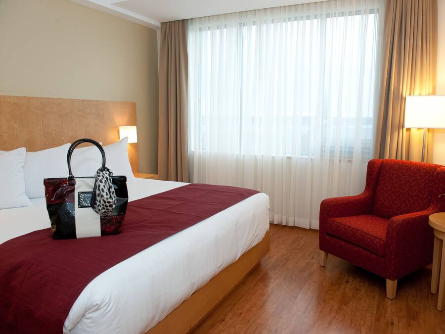A hotel room with a bed, a red armchair, a small table with a vase of flowers, and a window with curtains.