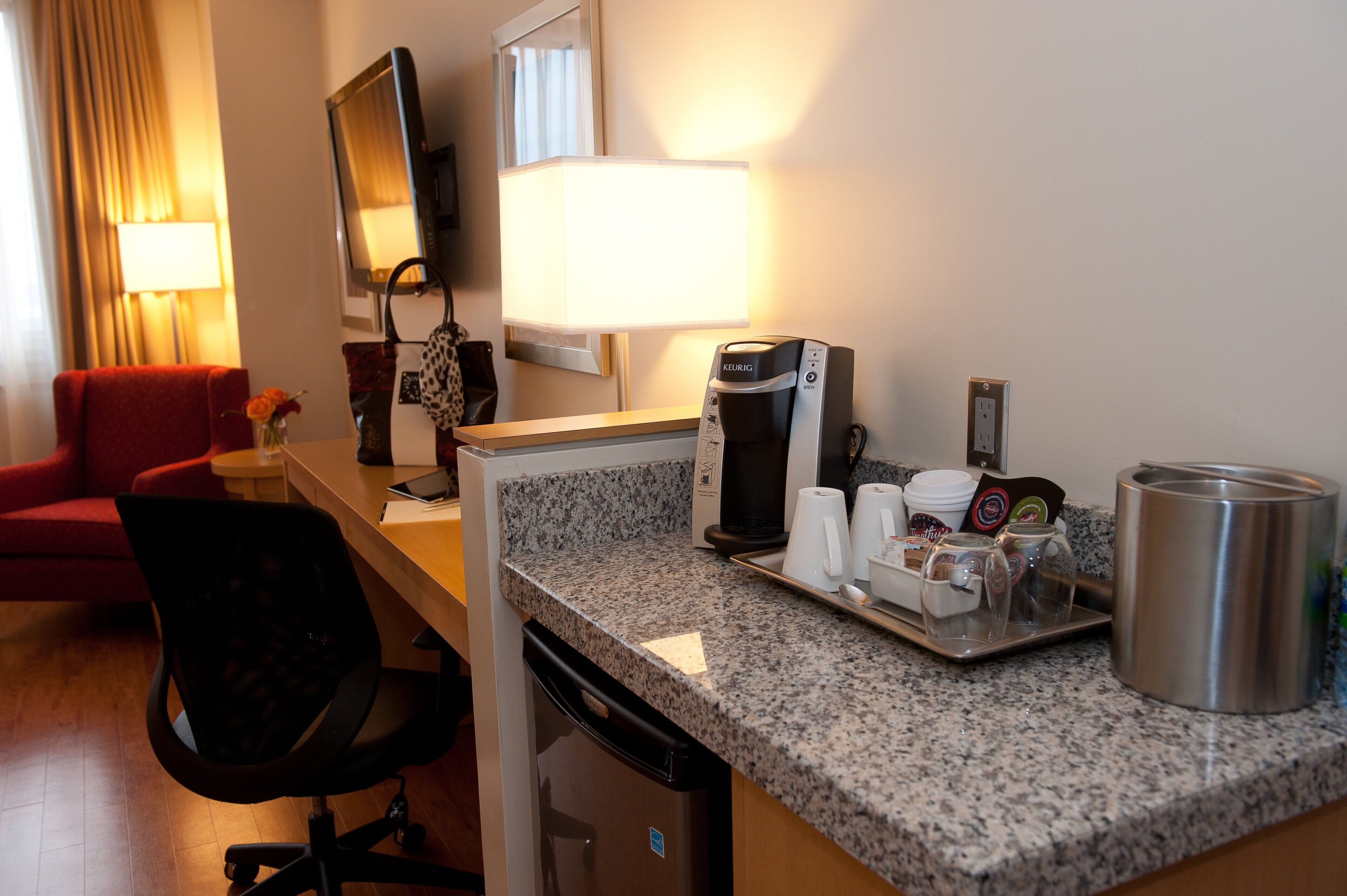 Habitación de hotel con encimera de granito, cafetera, hervidor de agua, bandeja con tazas y una taza, una silla roja, un escritorio pequeño, una lámpara y un televisor.