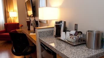 Habitación de hotel con encimera de granito, cafetera, hervidor de agua, bandeja con tazas y una taza, una silla roja, un escritorio pequeño, una lámpara y un televisor.