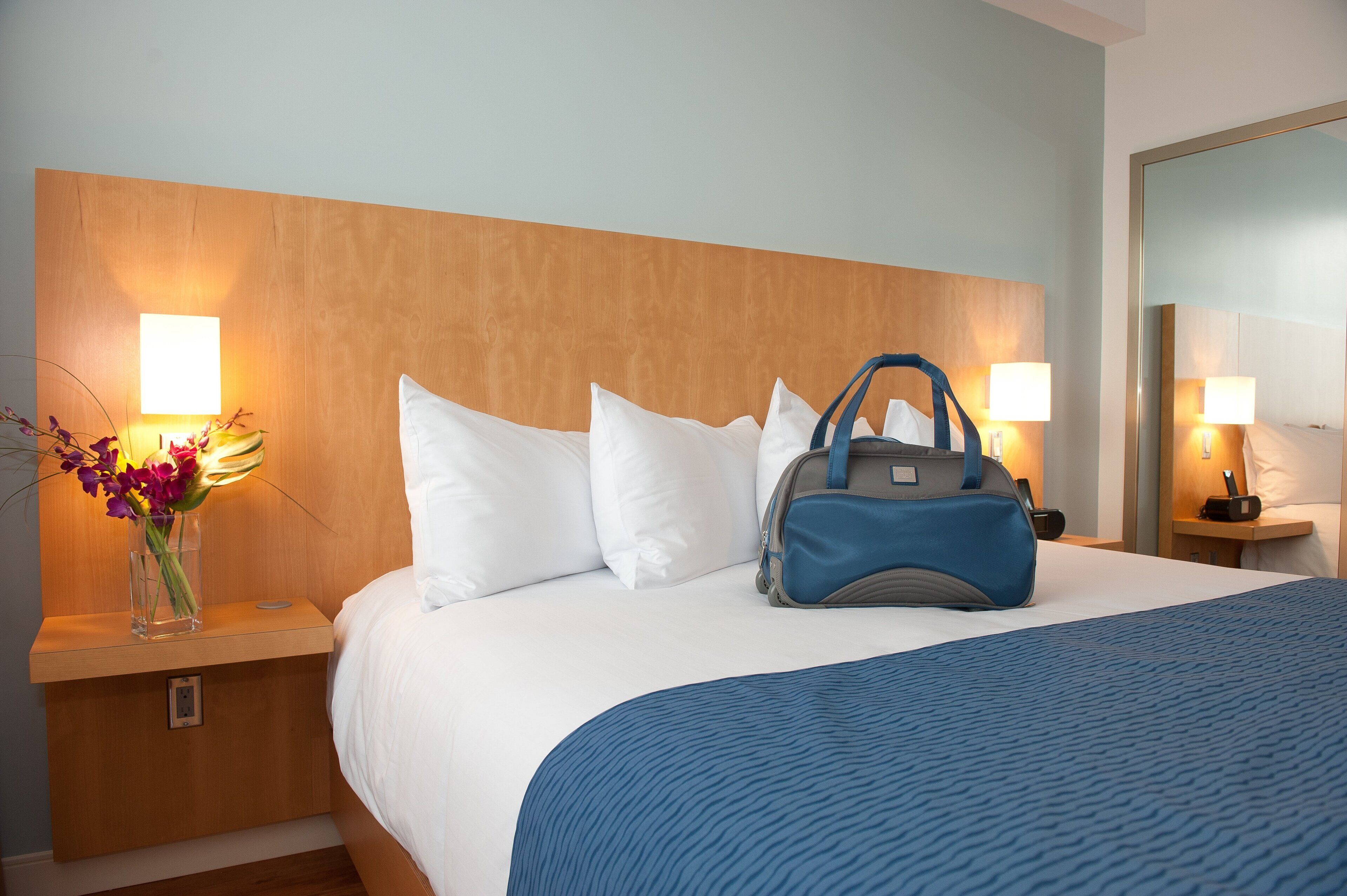 A hotel room with a bed, white pillows, a blue blanket, a wooden headboard, bedside lamps, a nightstand with a vase of flowers, and a blue bag on the bed.
