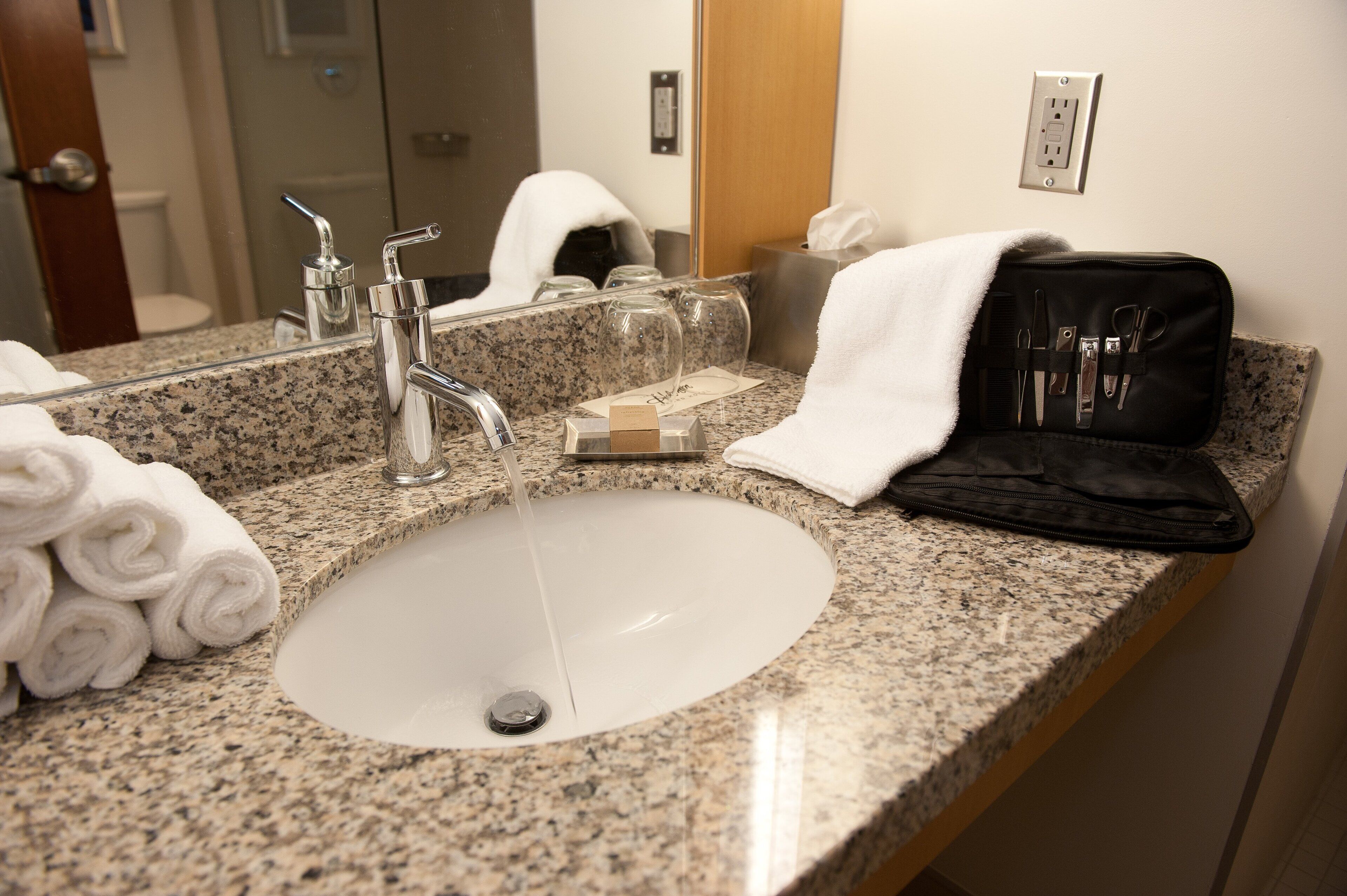 A bathroom with a granite countertop, a white sink, a mirror, and a black toiletry bag with scissors and a comb.